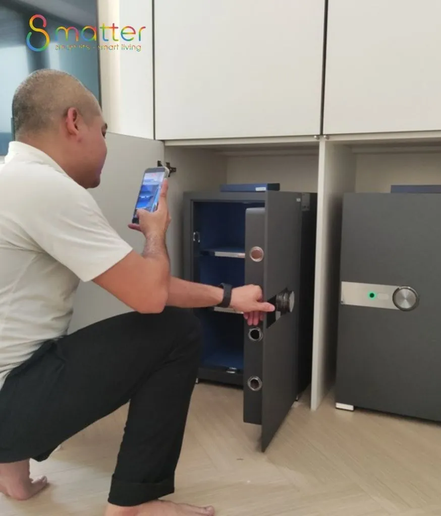 Người đàn ông ngồi mở két sắt xám lòng xanh, tay cầm điện thoại theo dõi