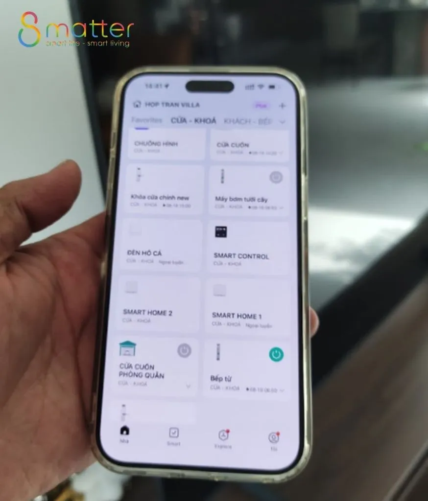 Bàn tay cầm iPhone mở app nhà thông minh liệt kê các thiết bị trong nhà