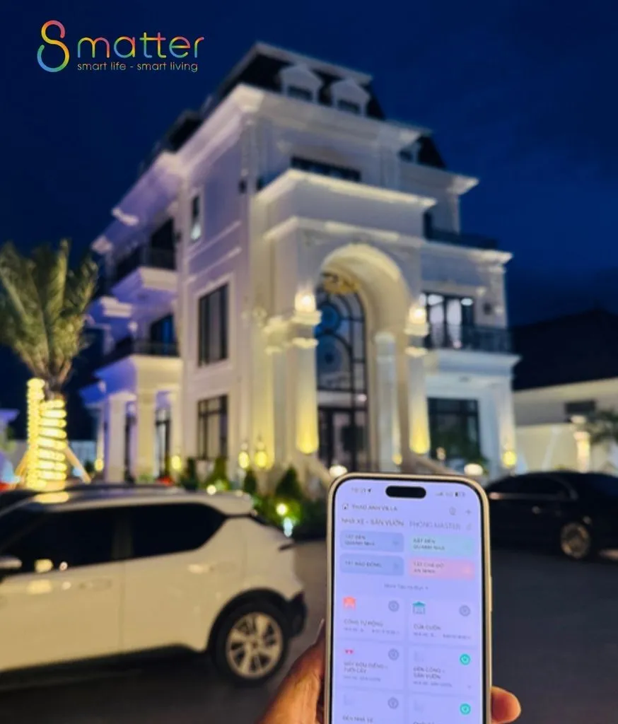Biệt thự trắng lên đèn về đêm, bàn tay cầm điện thoại mở app