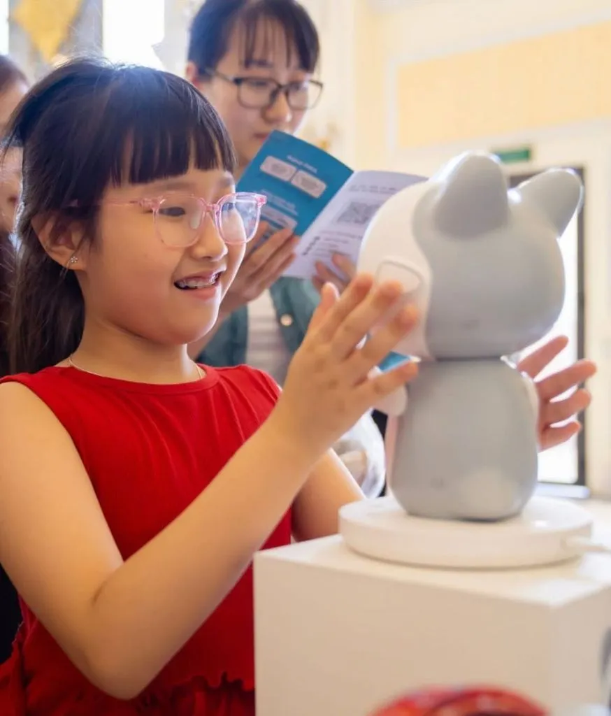Bé gái cầm tờ rơi giới thiệu, phía sau là robot Pika đặt trên bục trưng bày