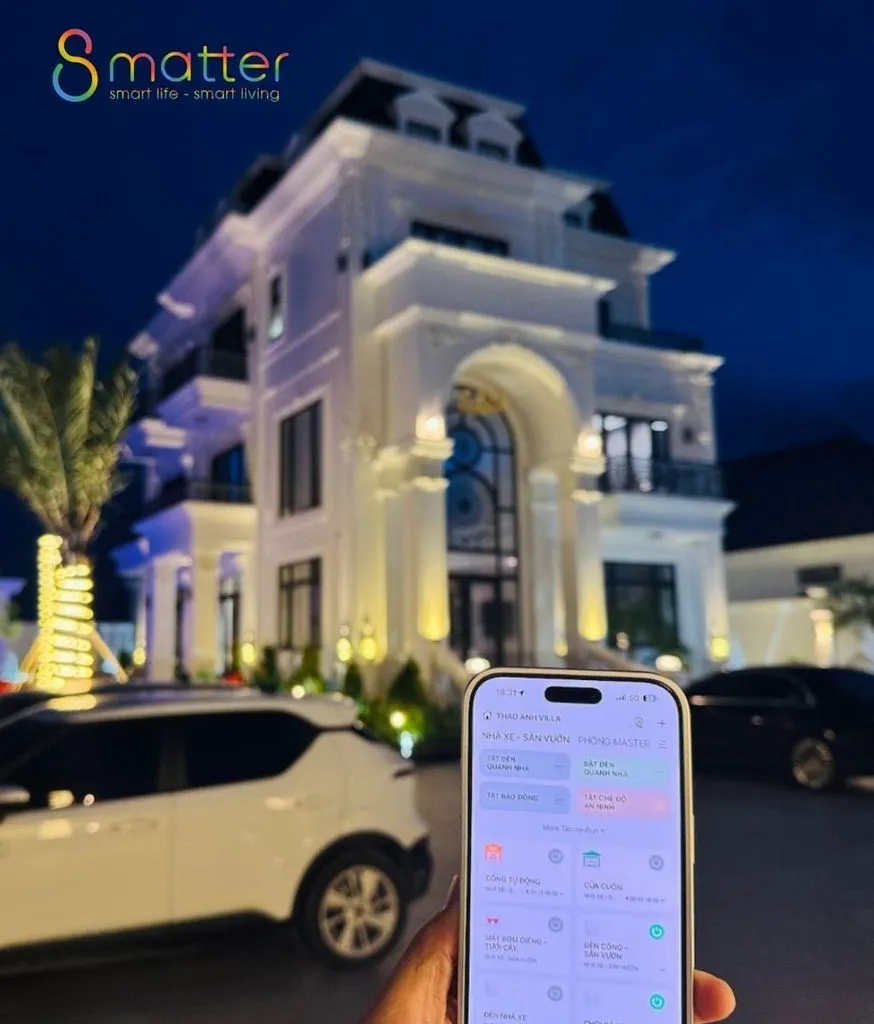 Căn biệt thự chụp ban đêm, phía trước là chiếc điện thoại mở ứng dụng điều khiển