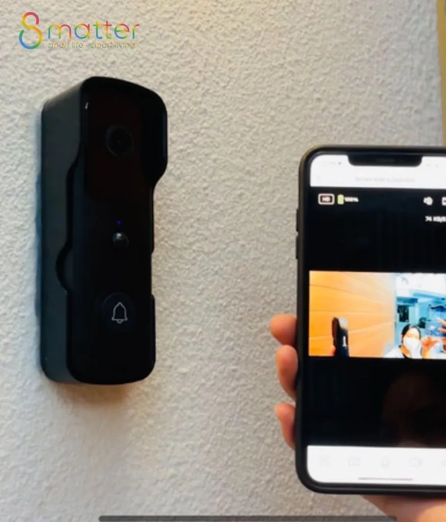 Chuông cửa camera đen gắn tường trắng, bàn tay cầm điện thoại hiển thị hình ảnh