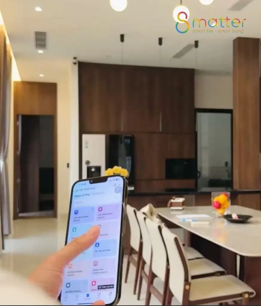 Bàn tay cầm điện thoại mở ứng dụng điều khiển, phía sau là phòng ăn và bếp