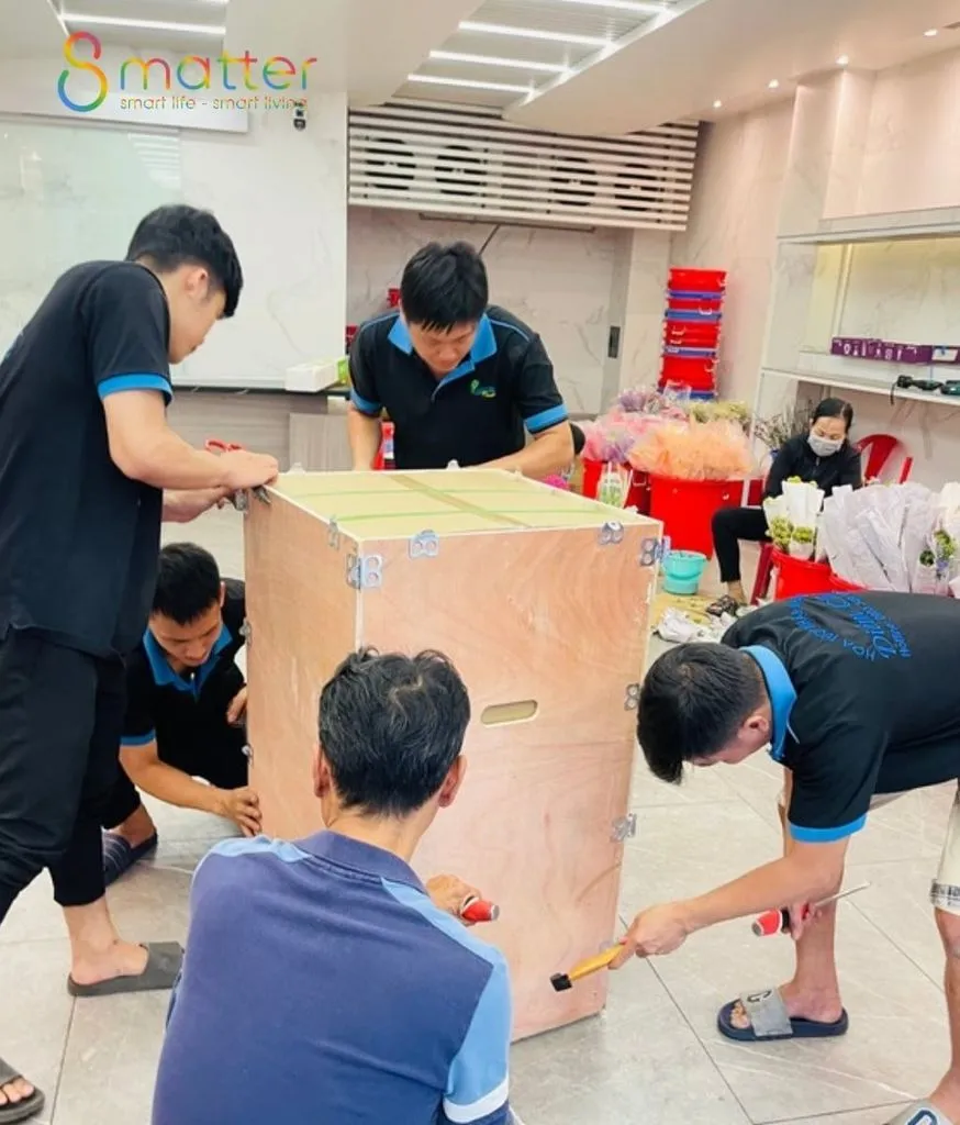 Nhóm nhân viên áo đen viền xanh đang đóng nẹp thùng gỗ lớn giữa cửa hàng