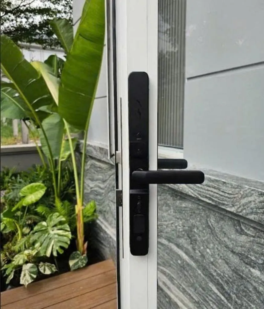 Khoá đen trên cửa nhôm trắng, cây chuối cảnh và bồn cây phía trước