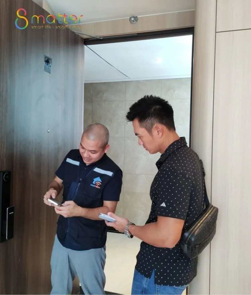 Hai người đàn ông đứng trước cửa gỗ, kỹ thuật viên áo xanh cầm điện thoại