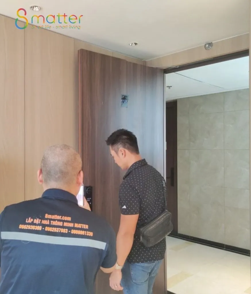 Kỹ thuật viên áo xanh in thông tin 8matter cùng khách nam xem cánh cửa gỗ trong sảnh căn hộ