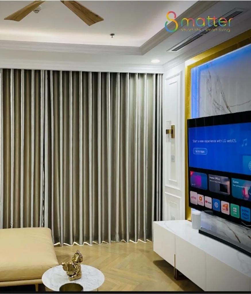 Phòng khách với rèm vải buông kín, tivi treo tường đang bật và bộ sofa trắng
