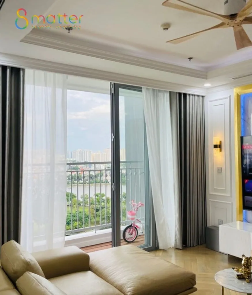 Phòng khách chung cư có ban công kính, rèm xám và voan trắng, sofa be và quạt trần