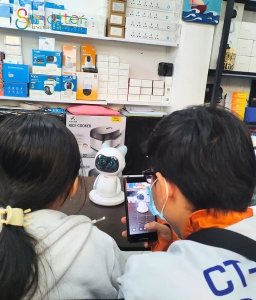 Hai người cúi xem robot Pika trắng đặt trên quầy cửa hàng điện