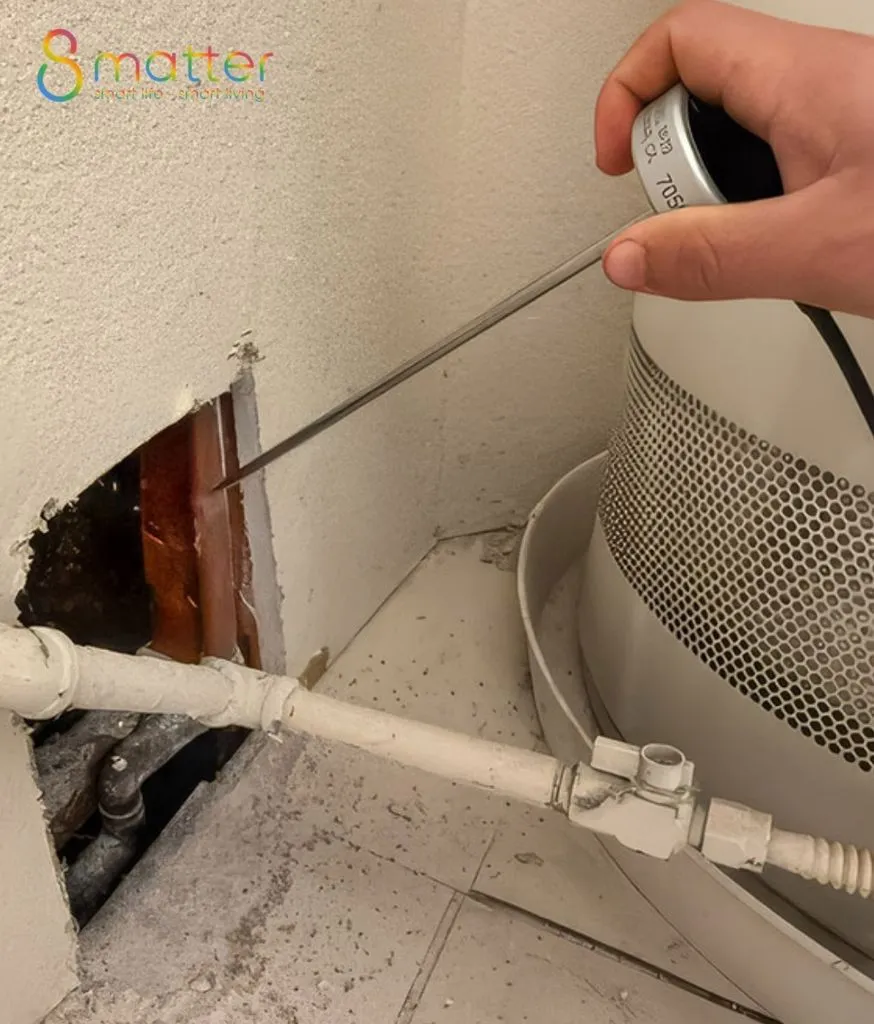 Bàn tay xịt bình foam vào lỗ đục trên tường trắng cạnh ống nước và thiết