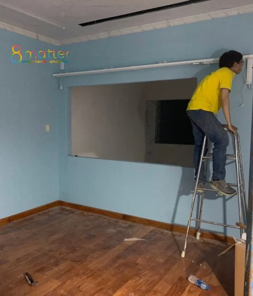 Người mặc áo vàng đứng thang thi công thiết bị trên tường phòng sơn xanh