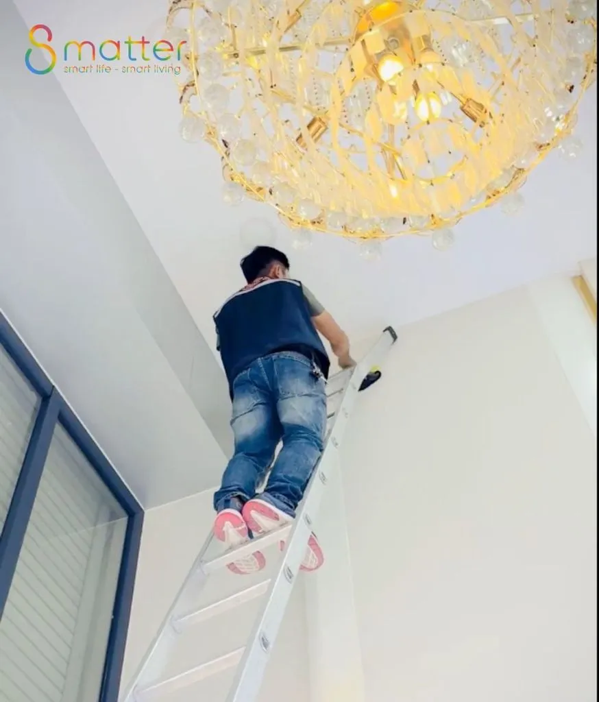 Thợ thi công đứng trên thang gấp lắp thiết bị trên trần nhà cạnh đèn chùm